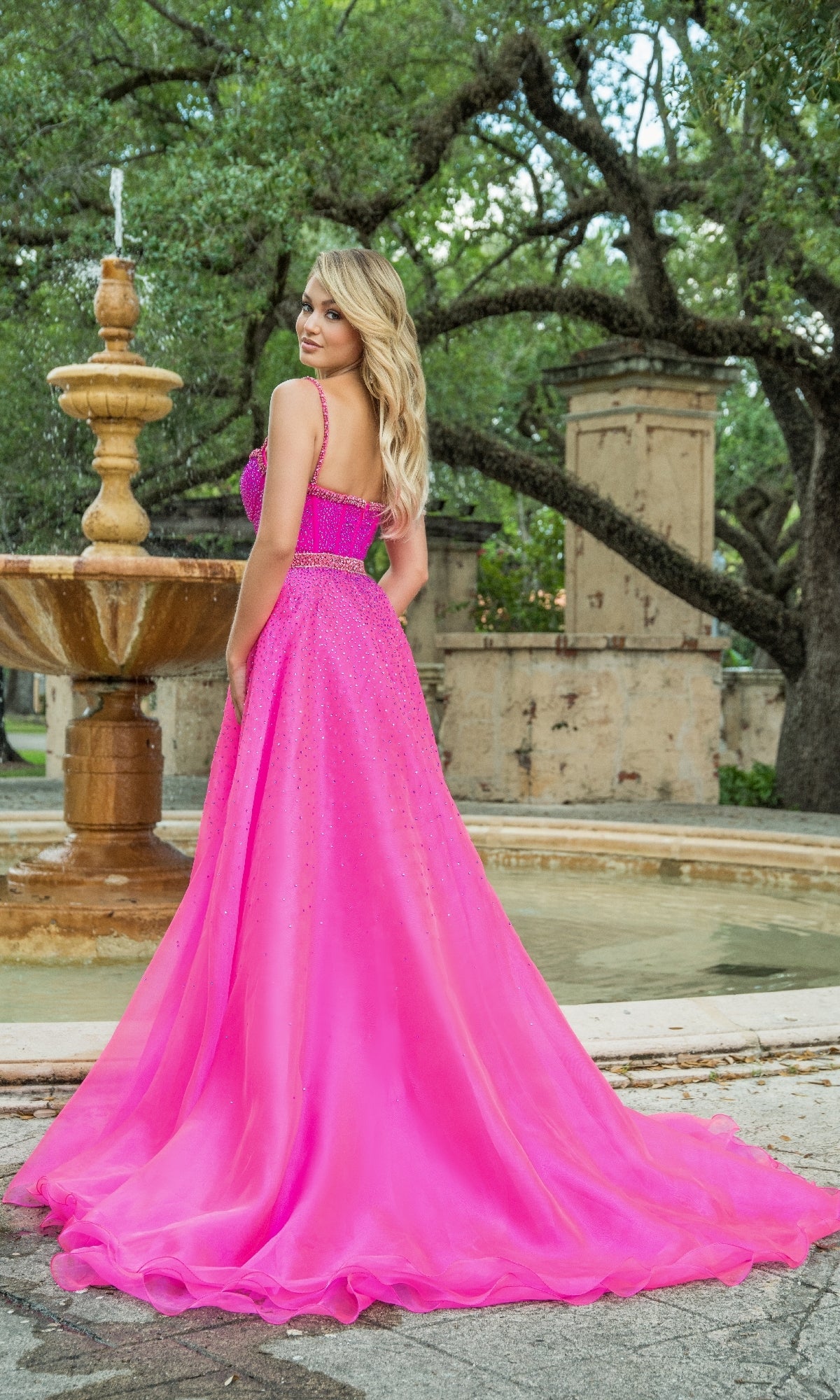 A woman in the Long Formal Dress 28581 by Ava Presley, featuring a bright pink corset bodice, stands before a stone fountain with trees and old walls providing a charming outdoor backdrop.